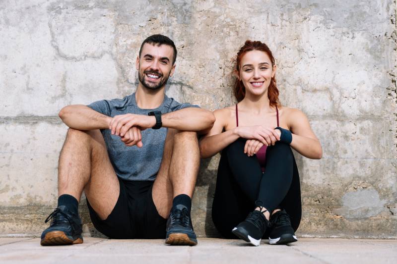 Exhausted young adult couple resting in the floor after running together in the park. Affectionate satisfied athletic male and female sitting in the grass in sportswear training and workout outside. 