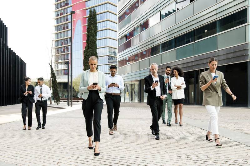 Group of confident business people walking the street using their phones. Diverse team of focused businesswoman and businessman walking outside while texting on their smartphones.