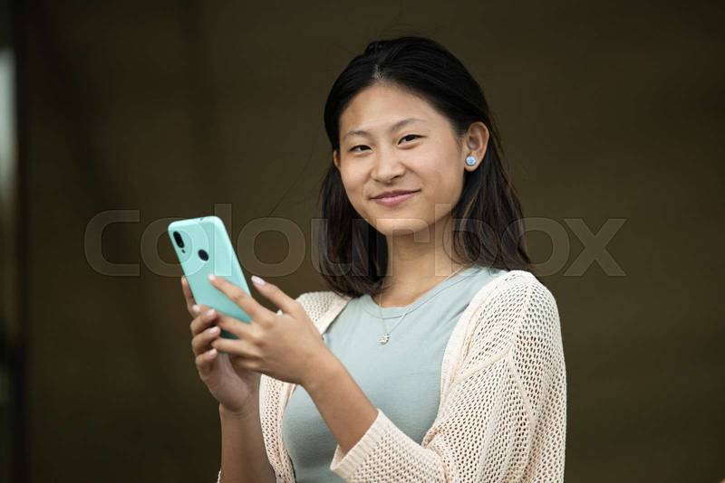 Beautiful cheerful young lady looking at camera texting and standing outside.Joyful carefree woman staring at camera in the street while using her phone.