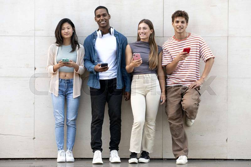 Diverse group of young people texting leaning against a wall looking at camera. Happy group of millennials friends using their phones standing in the street staring at the camera.