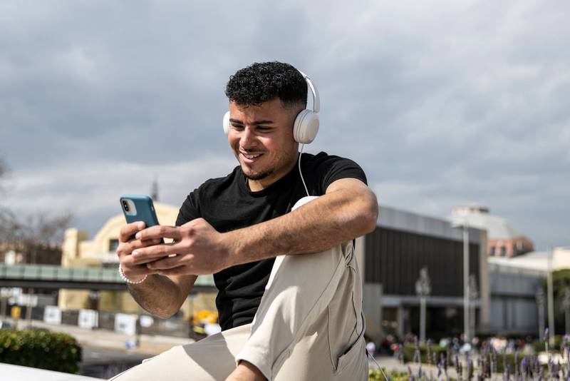 Young handsome man sitting on bank relaxed and texting with her phone using headphones. Satisfied guy smiling and using an smartphone sitting in the street listening to music.