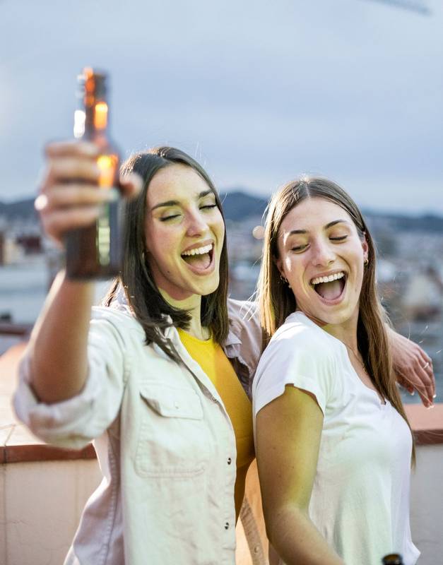Beautiful and cheerful couple dancing in a rooftop party while drinking beer. Two happy women singing together and having fun in a terrace party holding a beer bottle.