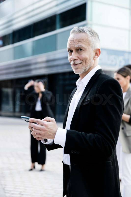 Side view of a mature handsome businessman smiling and looking at camera in the street while using his phone. Confident senior executive in black suite staring at camera.