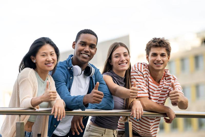 Beautiful group of friends satisfied and looking at camera leaning on a balcony. Diverse group of cheerful and hopeful millennial people showing thumb up.