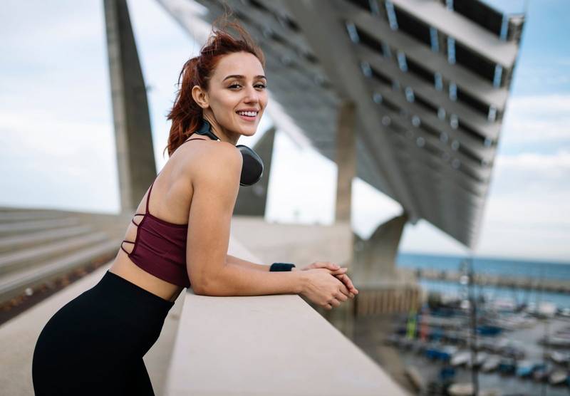 Young smiling fit woman in sportswear resting wearing outside headphones looking at camera. Athletic female listening music taking a breath during a training workout leaning on wall after exercising.