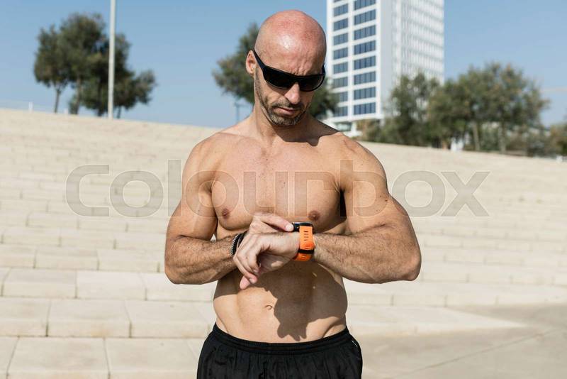 Fit man checking his watch shirtless with sunglasses outdoors. Handsome sport male looking at a smartwatch wearing black shorts in the street.