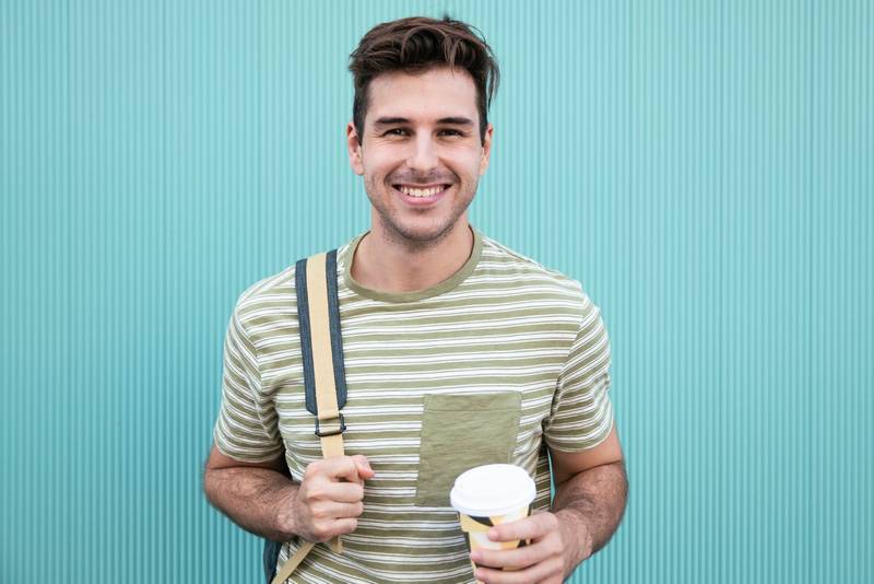 Portrait of handsome happy student smiling and drinking a take away coffee at a green wall.