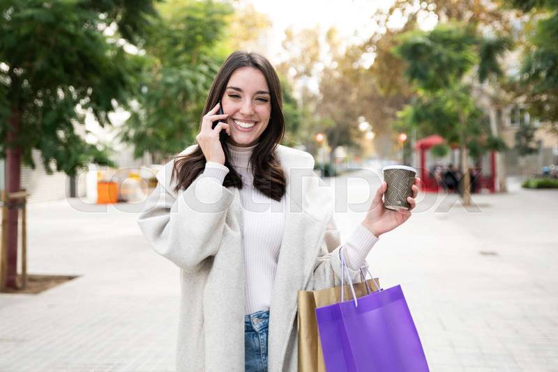 Portrait of beautiful young woman with shopping bags looking at camera with take away coffee and calling with cell telephone 