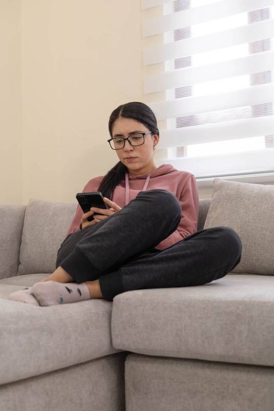 Casual young woman in pink hoodie and glasses sitting on sofa, smiling while using her smartphone