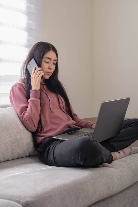Concentrated woman in a pink hoodie sitting on a couch and typing on her laptop at home, working