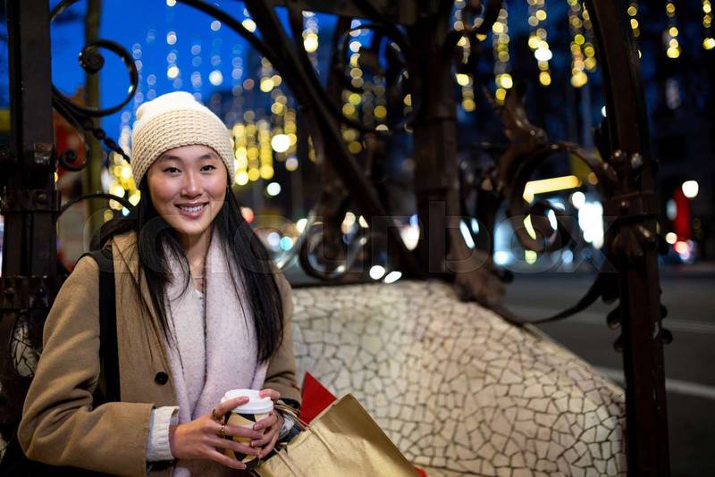 Joyful young woman shopping at evening looking at camera. Cheerful female holding a coffee standing at night in the street in winter.