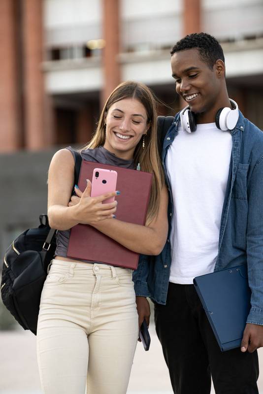 Multiracial couple standing and looking at a phone call in campus. Two young students taking a selfie smiling in the street.