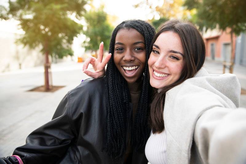 Two young multicultural friends taking selfie and having fun together - Caucasian and American Afro female frineds enjoying together.