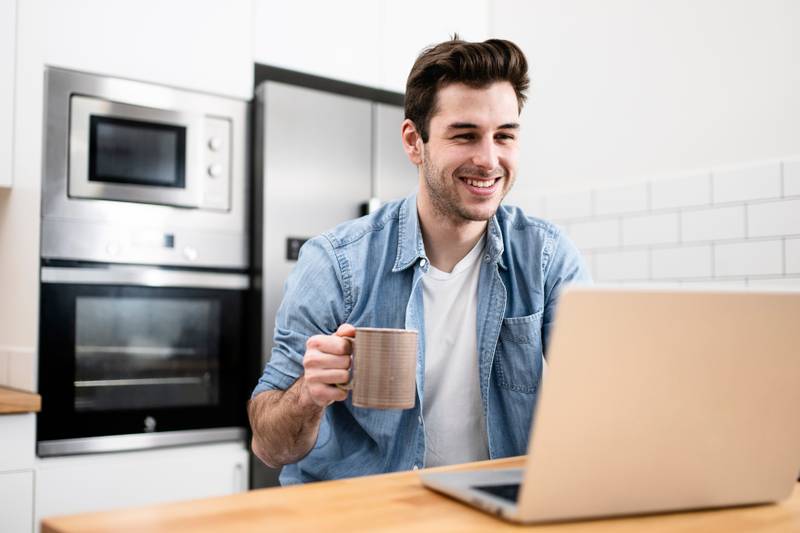 Handsome young man working with a laptop and smiling at the kitchen with a cup of tea - Cheerful, success and business concept