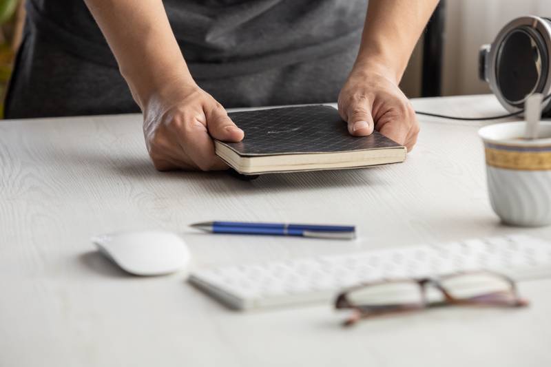 person holding a black notebook, desk with glasses, pen, cup of coffee, computer, technology