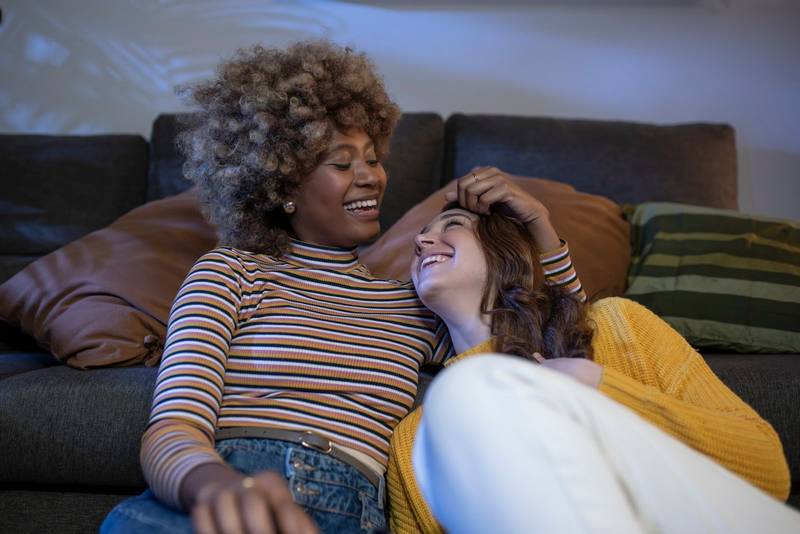 Multiracial lesbian couple hugging and laughing and relaxing in a living room. Smiling gay females couple having fun at home embracing each other.