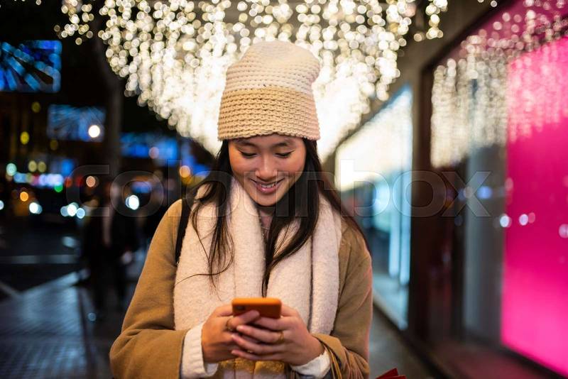 Young smiling girl texting in the street at night. Happy woman wearing warm hat using her phone in winter.