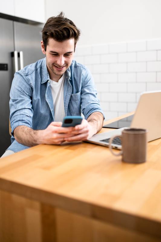 Handsome young man working with a laptop and smiling at the kitchen with a cup of tea - Cheerful, success and business concept