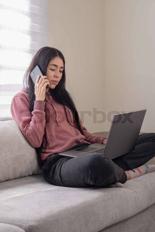 Concentrated woman in a pink hoodie sitting on a couch and typing on her laptop at home, working