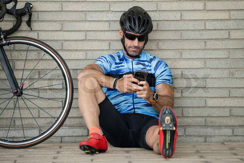 Fit happy cylcist man checking his phone sitting on the street floor. Relaxed smiling athletic bike rider male watching his smartphone resting against a wall in the city.