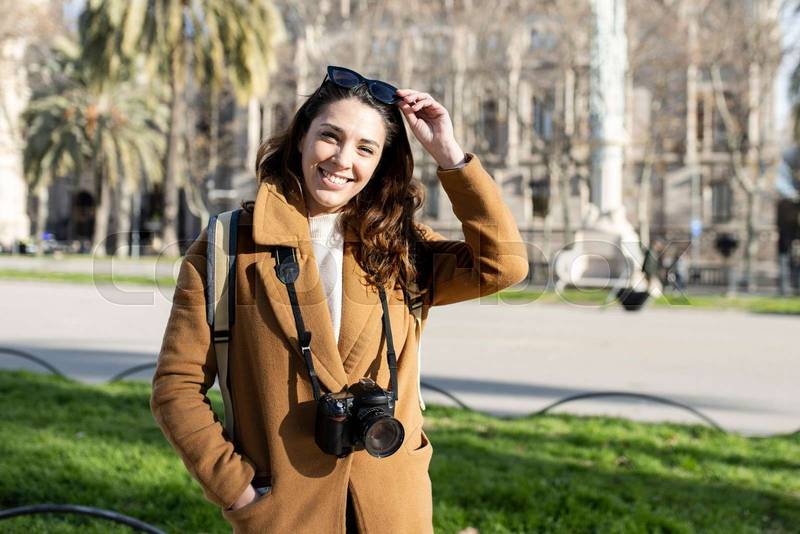 Smiling young female photographer on city street. Positive young female tourist with sunglasses wearing brown coat standing with hands in pockets and photo camera around neck.