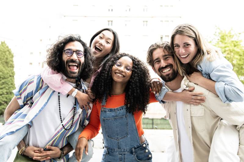 Multiracial group of friends giving piggyback ride and having fun together looking at camera.Diverse young women and men hugging each other laughing and carefree staring at camera outside.