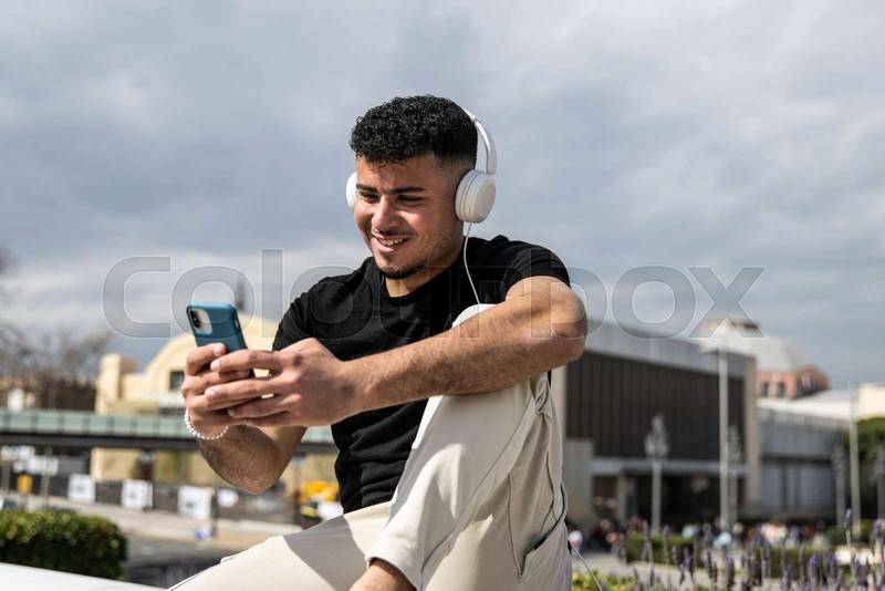 Young handsome man sitting on bank relaxed and texting with her phone using headphones. Satisfied guy smiling and using an smartphone sitting in the street listening to music.