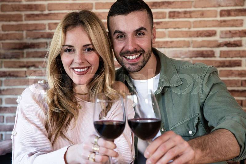  Cheerful male and female drinking with glasses at home. Joyful young adult couple toasting with wine and looking at camera.