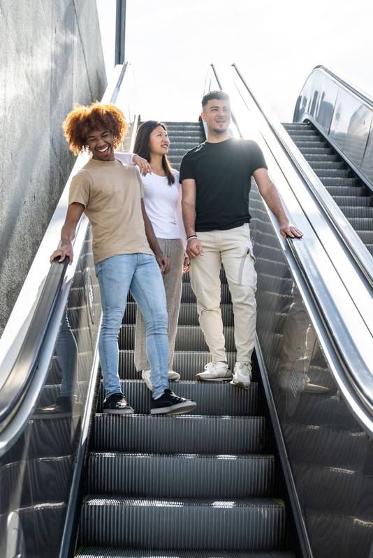 Multiracial group of friends laughing and going down an outdoor escalator. Three happy young people smiling and looking relaxed walking down of stairs in the street.