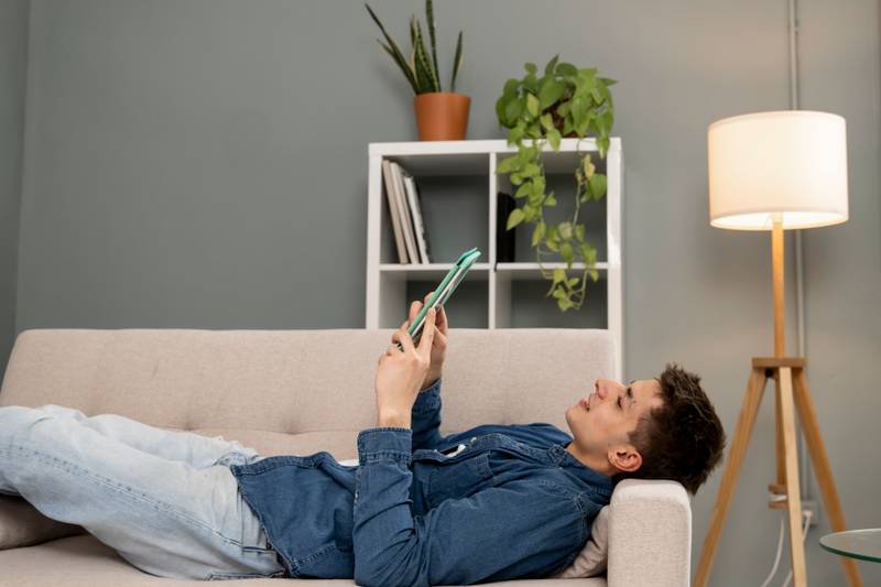 Relaxed young carefree man watching video with a tablet at home. Handsome male resting using with touchpad while sitting on a couch.