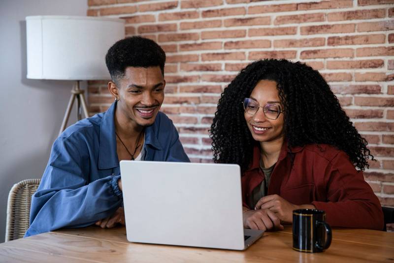 Handsome young couple using laptop together while sitting at home. Smiling friends looking at computer and laughing at cozy home office.
