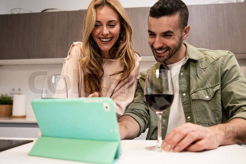 Happy joyful man and woman on a video call having a glass of wine. Young adult couple using together a tablet from home in a modern kitchen.