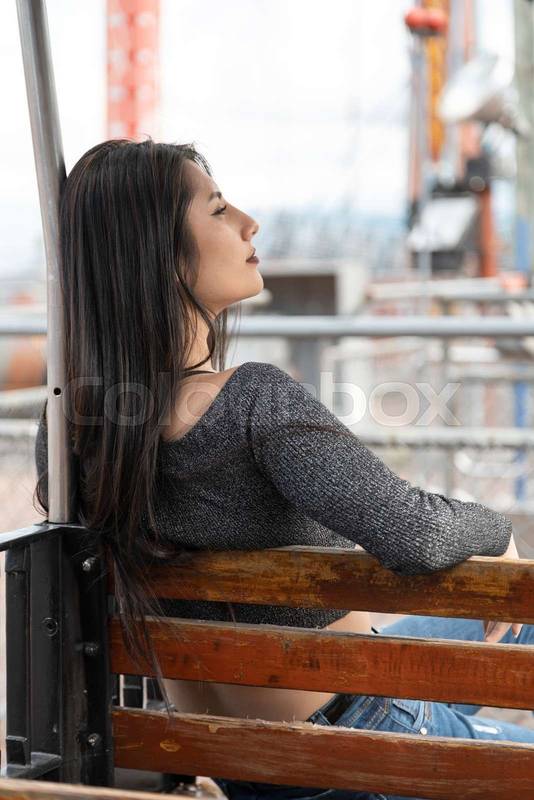 sitting on a wooden bench with her face in profile, relaxed resting in a park outdoors, lifestyle