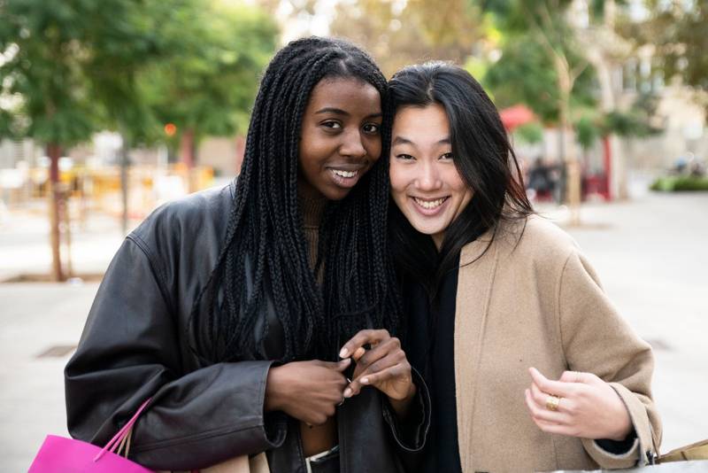 Portrait of two beautiful young women looking at camera and holding shopping bags. Smiling multiethnic friends together in the street