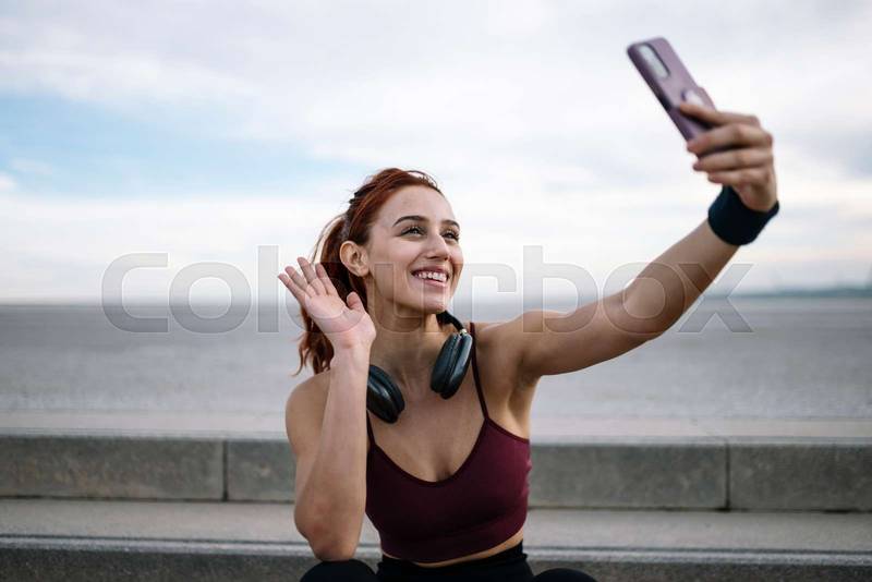 Joyful sports woman taking a selfie wearing headphones sitting outside. Young adult fit athletic female in sportswear taking a picture with smartphone