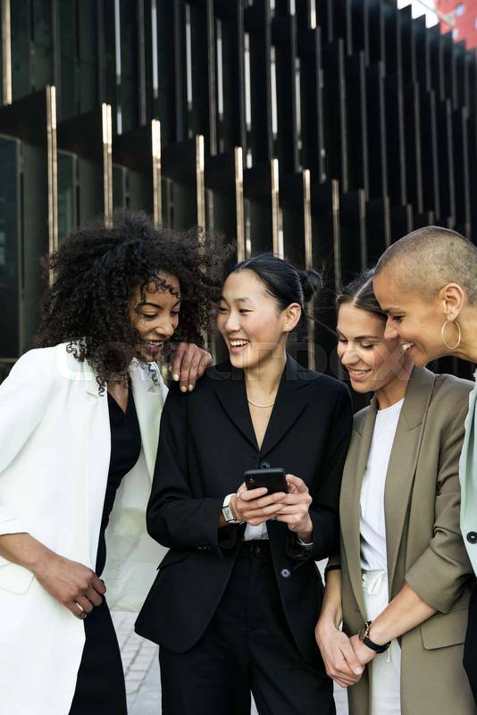 Diverse businesswomen group looking a phone and laughing standing in the street. Casual meeting between young females executives checking their smartphone outisde.